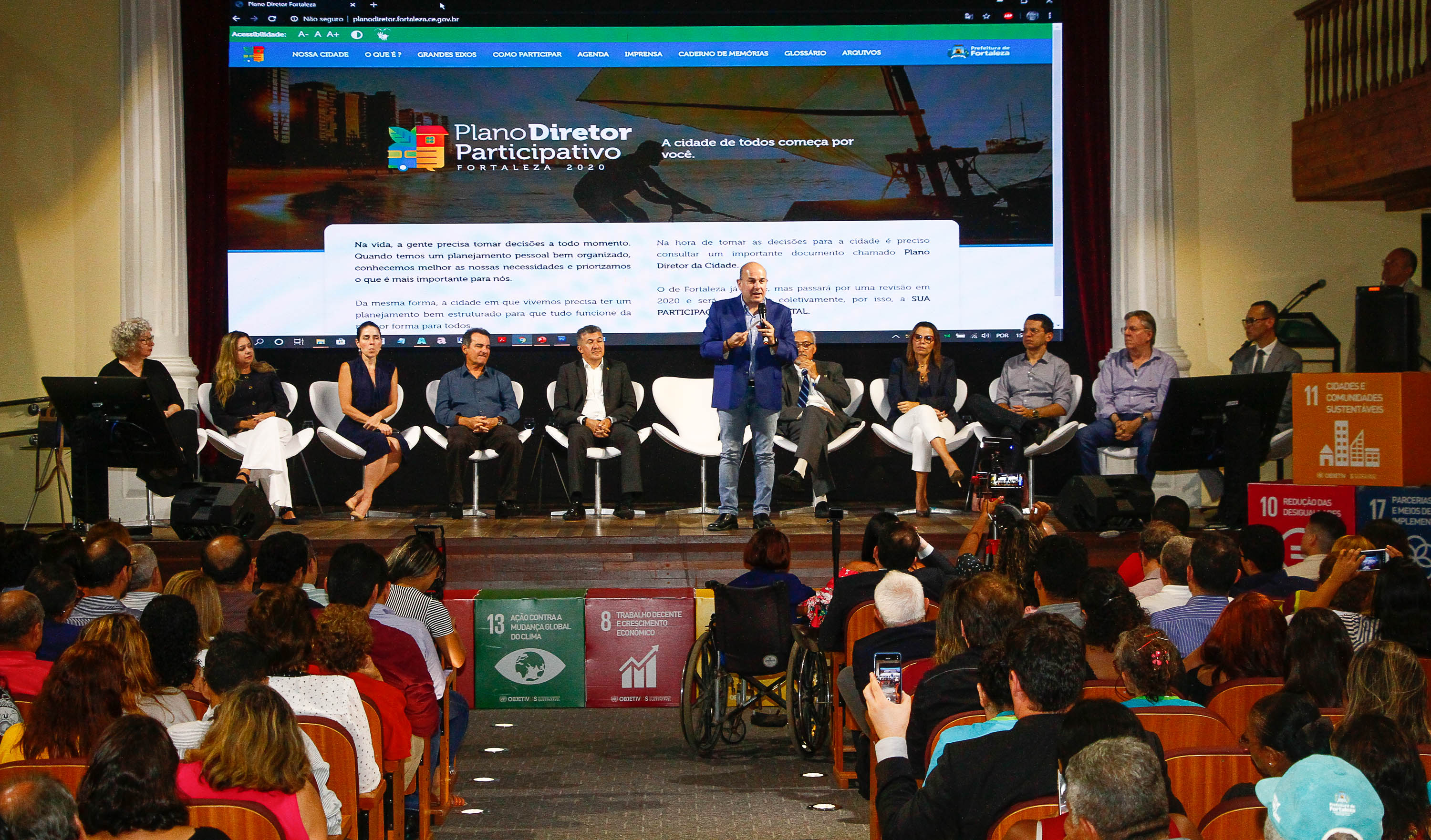 prefeito em cima de palco falando ao microfone, pessoas sentadas atrás e plateia sentada à frente assistindo